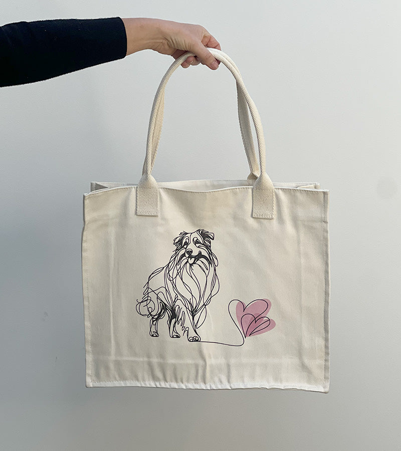 Tote bag with a Australian Shepherd dog illustration and pink hearts held by a hand against a plain background