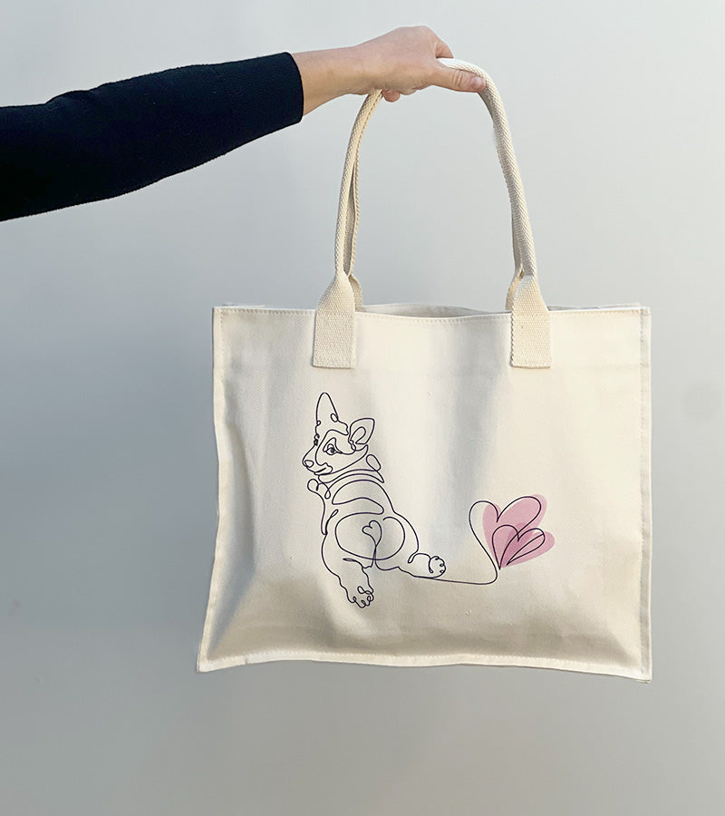 Tote bag with a Corgi dog illustration and pink hearts held by a hand against a plain background