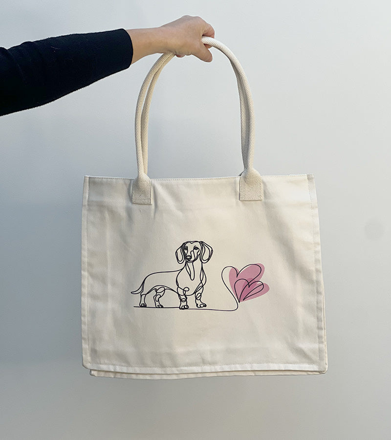Tote bag with a Dachshund dog illustration and pink hearts held by a hand against a plain background