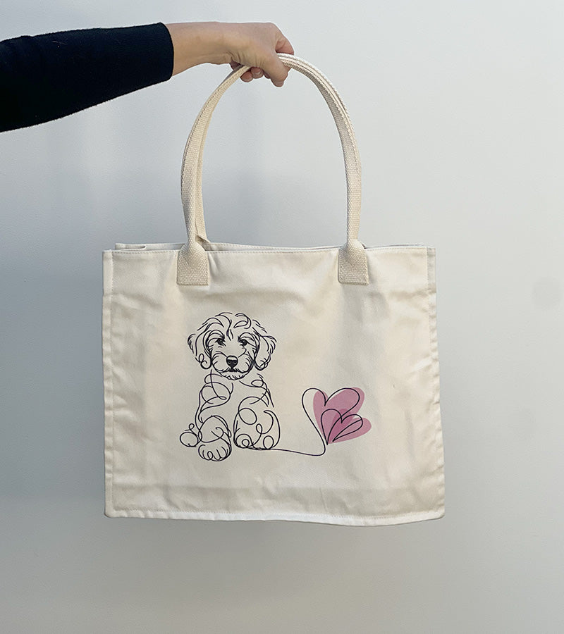 Tote bag with a Doodle dog illustration and pink hearts held by a hand against a plain background