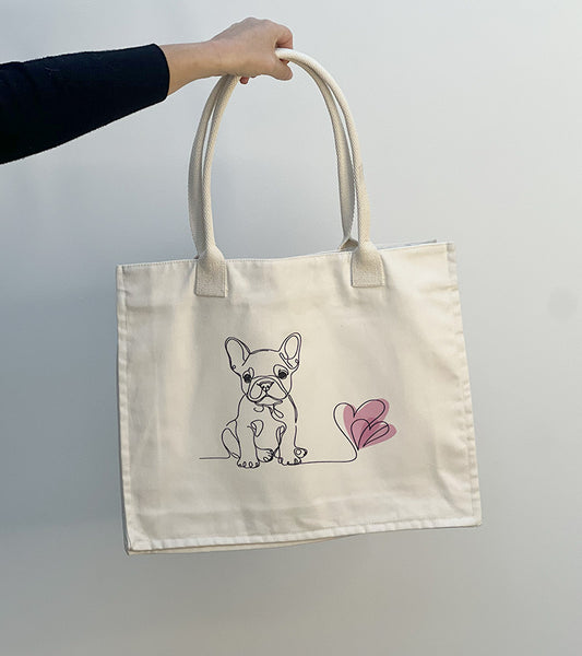 Tote bag with a French Bulldog illustration and pink hearts held by a hand against a plain background