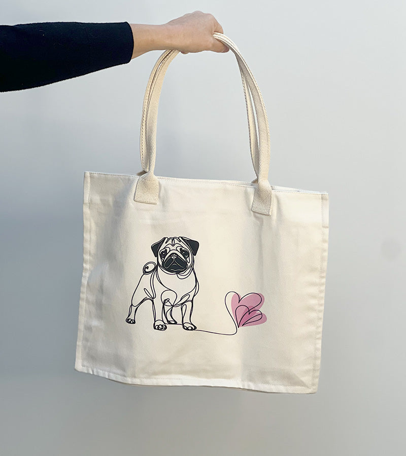 Tote bag with a Pug dog illustration and pink hearts held by a hand against a plain background