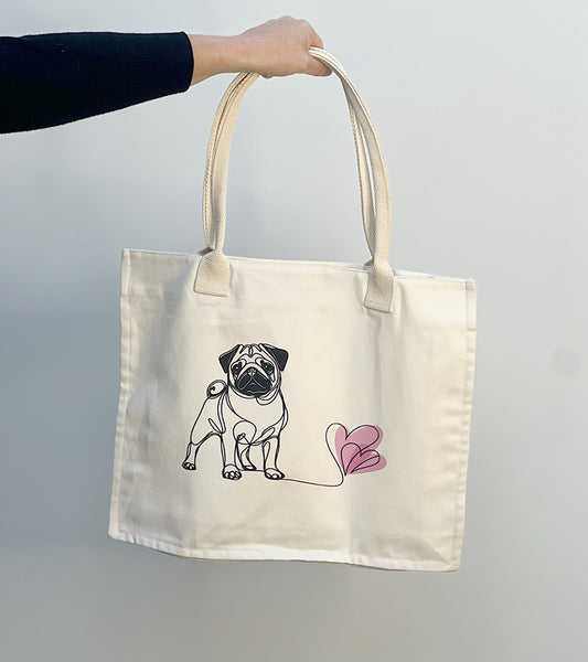 Tote bag with a Pug dog illustration and pink hearts held by a hand against a plain background