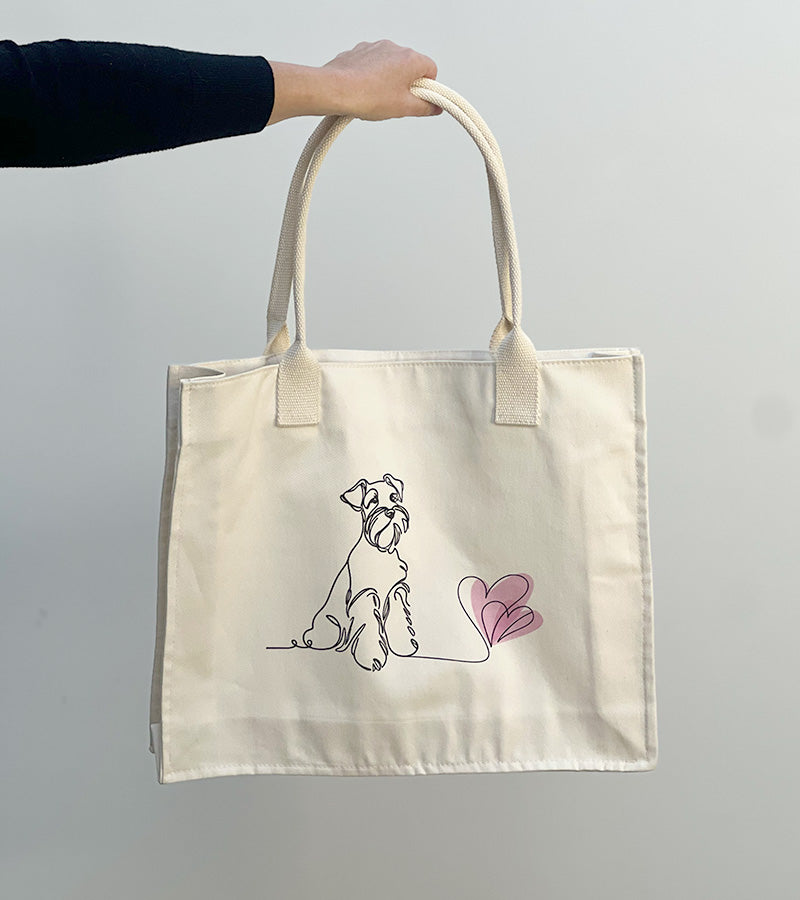 Tote bag with a Schnauzer dog illustration and pink hearts held by a hand against a plain background