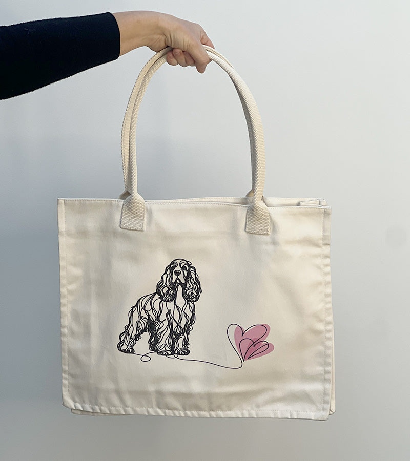 Tote bag with a Spaniel dog illustration and pink hearts held by a hand against a plain background