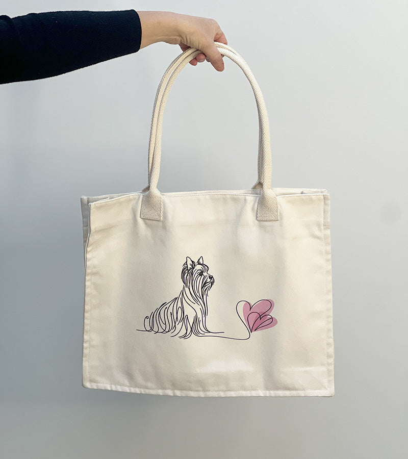 Tote bag with a Yorkie dog illustration and pink hearts held by a hand against a plain background